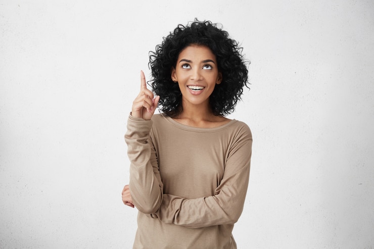 Free Photo look at that! indoor portrait of cheerful attractive casually dressed young female with curly hair pointing her index finger up, indication something interesting, having happy excited look