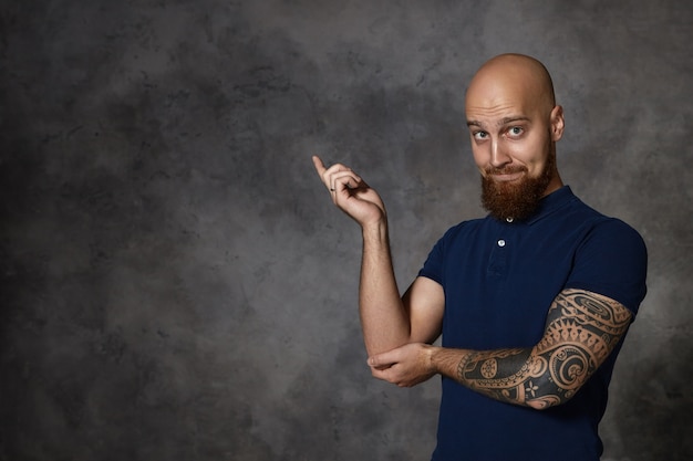 Free photo look at that! isolated funny bald bearded man with tattoo raising index fingre and pointing at left corner, expressing excitement or curiosity, raising eyebrows. body language