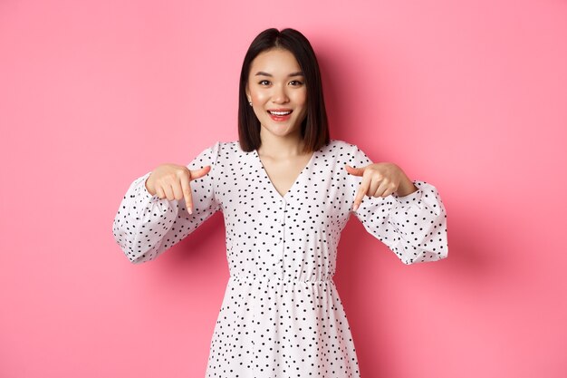 あっち見て。ドレスを着たかわいいアジアの女性、コピースペースで指を下に向けて、製品の割引を表示し、笑顔、ピンクの背景の上に立って