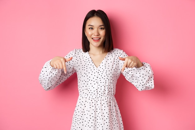 Посмотрите там. Симпатичная азиатская женщина в платье, указывая пальцами вниз на копировальное пространство, показывая скидку на продукт и улыбаясь, стоит на розовом фоне