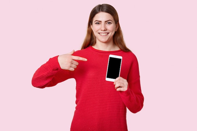 Look at this gadget. Happy smiling European young woman points at modern cell phone with empty screen for your promotional content