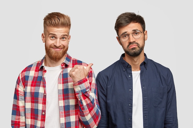 Look at this guy! Ginger bearded young man points with thumb at his fellow who has hesitant expression