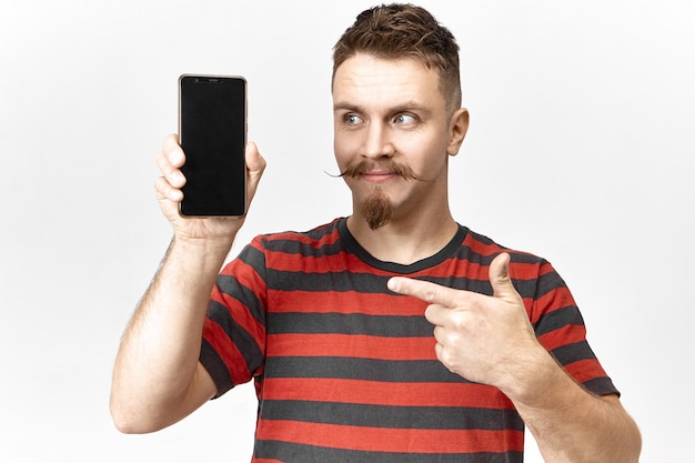 Look at this. Handsome emotional young salesman with stylish beard and mustache posing in studio with smart phone in his hand, pointing fore finger at blank copyspace screen, presenting gadget