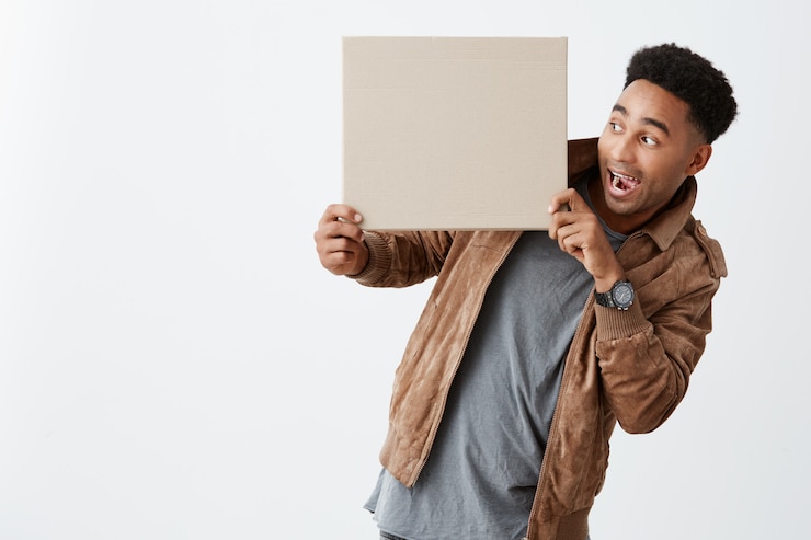 Free Photo look at this. isolated on white portrait of dark-skinned funny guy with afro hairstyle in casual winter clothes holding cardboard in hands, looking at it with opened mouth and shocked expression.
