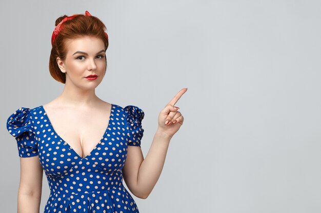 Look at this. Picture of gorgeous young Caucasian pin up woman wearing bright make up and low neck retro dress pointing fore finger, demonstrating advertising content on blank gray studio wall