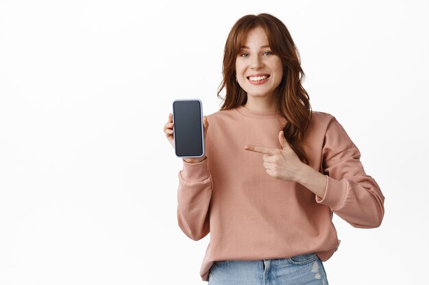Look at this. Smiling young woman pointing finger at smartphone blank screen, showing app or online shopping store, recommending download application, standing on white.