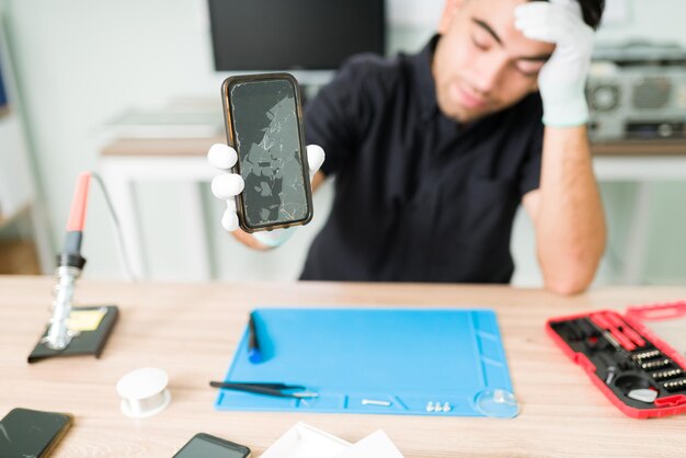 Look at this ugly smartphone. Upset male technician feeling worried while showing the cracked screen of a new smartphone