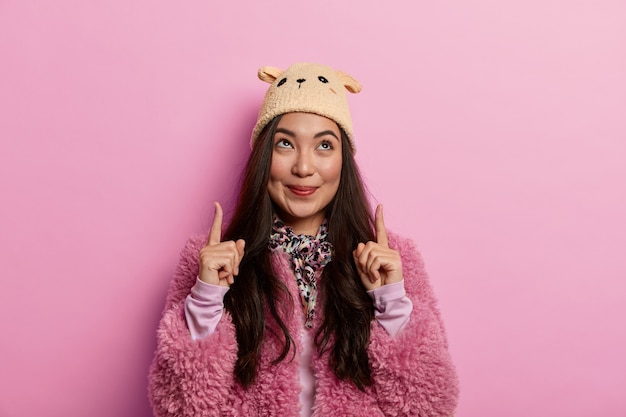 Look up. Attractive brunette woman points index fingers upwards, demontrates cool item with pleasant tender smile, wears pink coat