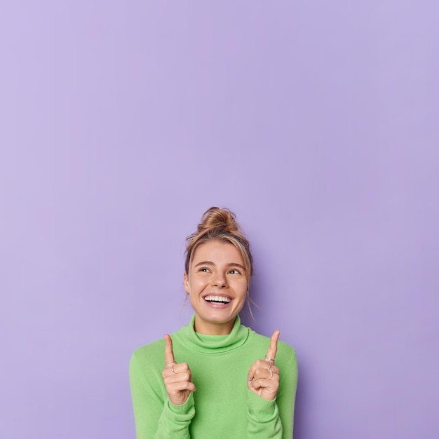 Бесплатное фото Посмотрите, какое хорошее предложение для вас. веселая оптимистичная женщина указывает на пространство для копирования выше и предлагает проверить это, демонстрируя свободное место для вашей рекламы, носит зеленую водолазку, изолированную на фиолетовой стене