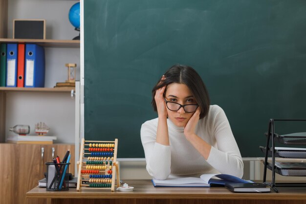 無料写真 教室で学校の道具をつけて机に座っている若い女教師の頭に手を置いてカメラを見て