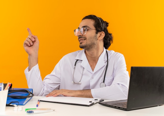 机に座っている聴診器で医療用ローブを着た医療用眼鏡をかけた若い男性医師の笑顔の側を見る