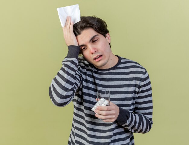 Бесплатное фото Глядя на бок усталого молодого больного человека, держащего салфетку с таблетками и положившего руку на голову, изолированную на оливково-зеленом фоне
