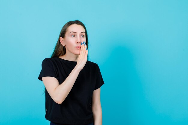 Looking away girl is trying say something by holding hand near mouth on blue background