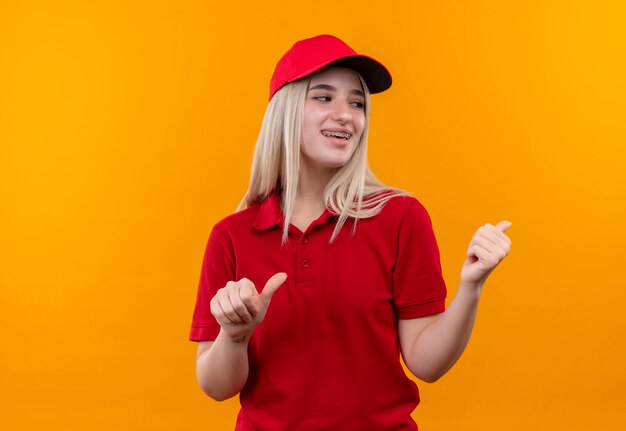 Looking at back smiling delivery young woman wearing red t-shirt and cap in dental brace points at back on isolated orange wall