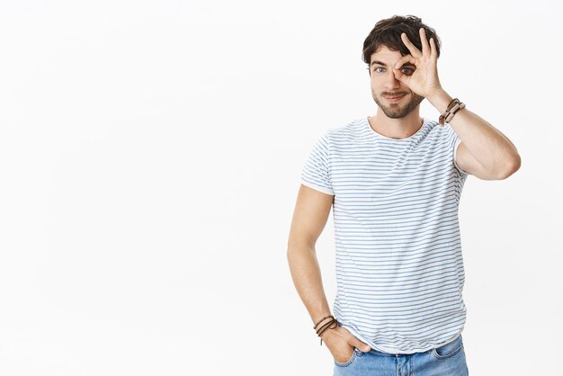 Looking on bright side is ok Portrait of goodlooking relaxed and friendly young masculine male with beard and blue eyes showing okay gesture on eye peeking through at camera with happy smile