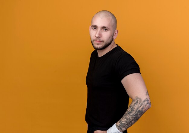 Looking at camera confident young sporty man wearing wristband putting hands on hip isolated on orange background with copy space