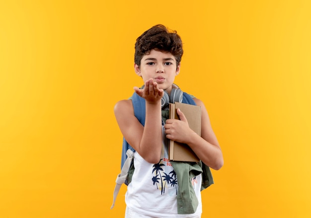 Looking at camera little school boy wearing back bag and headphones holding book and showing kiss gesture isolated on yellow background