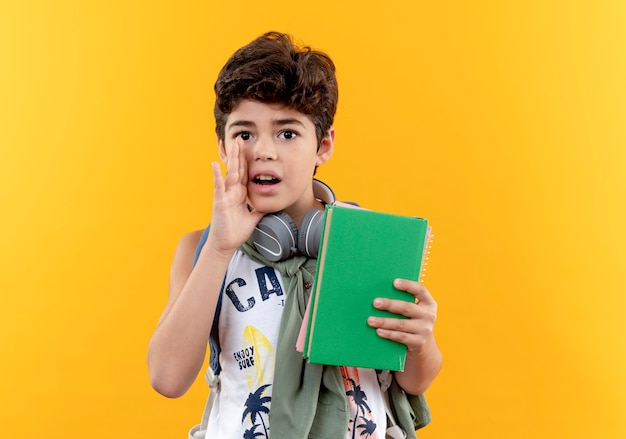 Looking at camera little school boy wearing back bag and headphones holding book and whisper isolated on yellow background