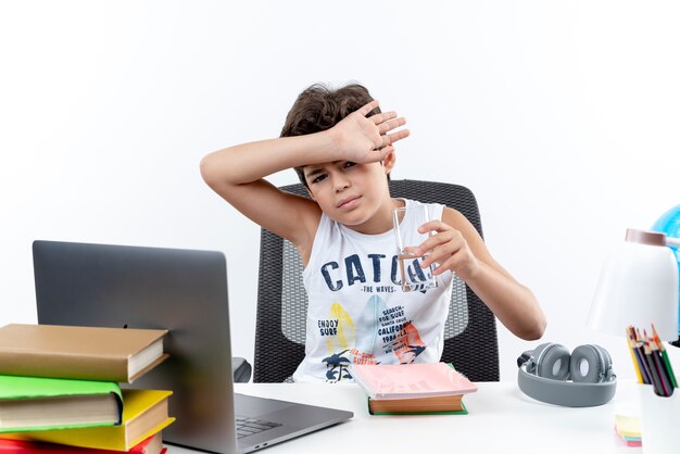 Looking at camera little schoolboy sitting at desk with school tools holding glass of water and covered forehead with wrist isolated on white background