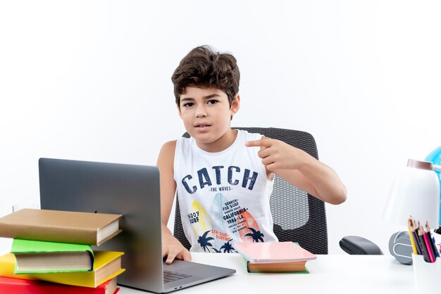 Looking at camera little schoolboy sitting at desk with school tools points at side isolated on white background with copy space