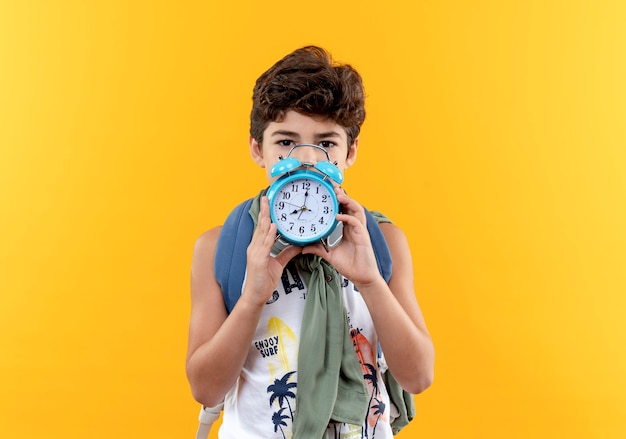 Looking at camera little schoolboy wearing back bag and headphones covered face with alarm clock isolated on yellow background