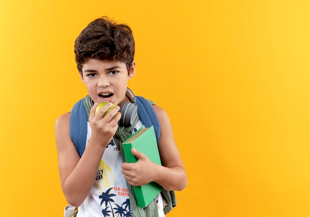 Looking at camera little schoolboy wearing back bag and headphones holding book and bites off apple isolated on yellow background