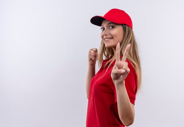 Looking at camera pleased young delivery girl wearing red uniform and cap showing peace gesture  isolated on white background with copy space