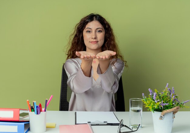 Looking at camera pleased young pretty female office worker sitting at desk with office tools holding out hands on camera isolated on olive background