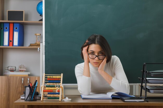 looking at camera putting hands on head young female teacher sitting at desk with school tools on in classroom