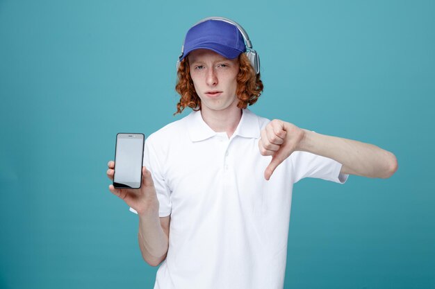 Глядя в камеру, показывая большие пальцы вниз, молодой красивый парень в кепке в наушниках держит телефон на синем фоне