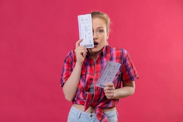 Looking at camera surprised young female traveler wearing red shirt covered eye with ticket on isolated pink wall