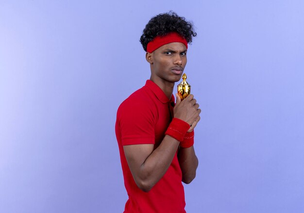 Looking at camera unpleased young afro-american sporty man wearing headband and wristband holding winner cup around chin isolated on blue background