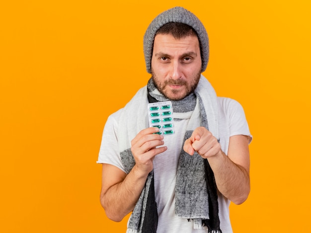 Looking at camera unpleased young ill man wearing winter hat and scarf holding pills and showing you gesture isolated on yellow background