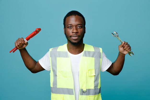 Looking at camera young african american builder in uniform holding open end wrench with gas wrench isolated on blue background
