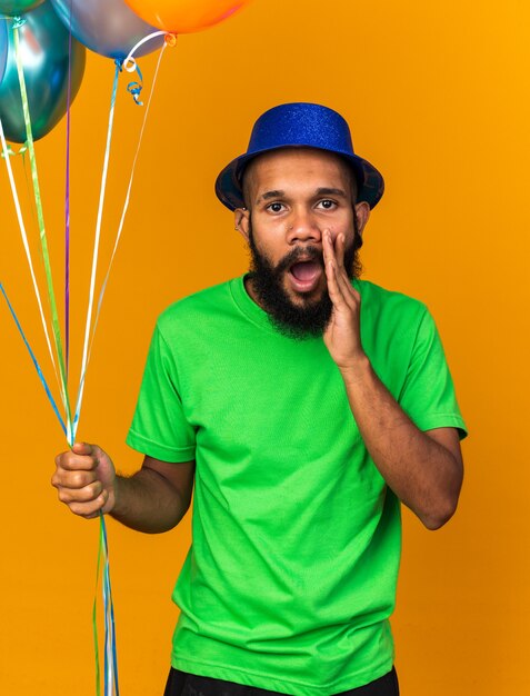 Looking camera young afro-american guy wearing party hat holding balloons calling someone 