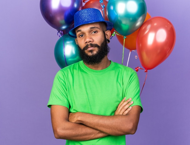 Глядя в камеру молодой афро-американский парень в партийной шляпе, стоящий перед скрещенными руками над воздушными шарами