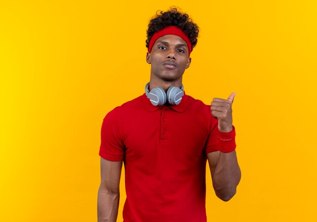 Looking at camera young afro-american sporty man wearing headband and wristband in headphones points at side isolated on yellow background with copy space