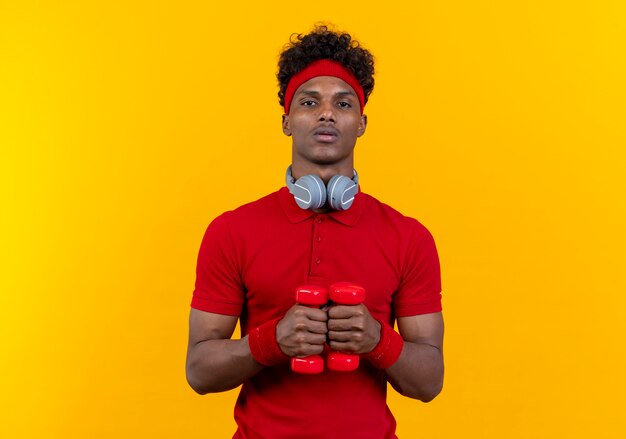 looking at camera young afro-american sporty man wearing headband and wristband holding dumbbells