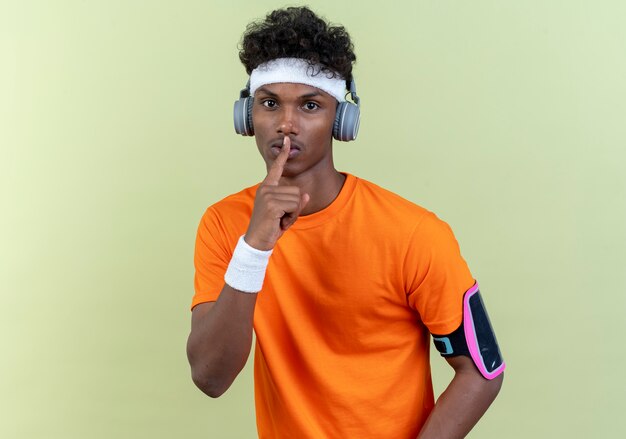 Looking at camera young afro-american sporty man wearing headband and wristband and phone arm band with headphones showing silence gesture isolated on green background