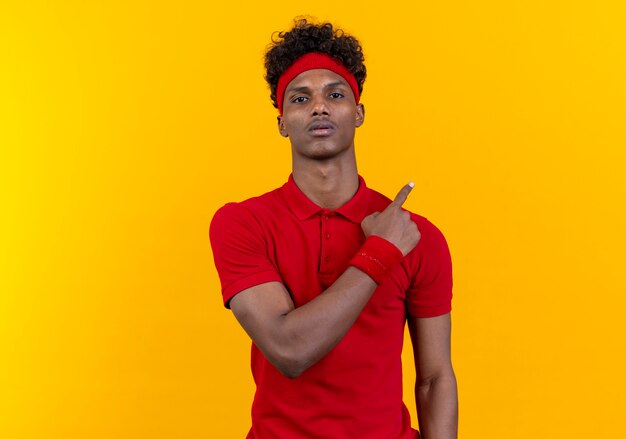 Looking at camera young afro-american sporty man wearing headband and wristband points at side isolated on yellow background with copy space