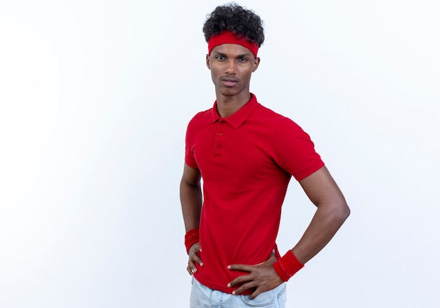 Looking at camera young afro-american sporty man wearing headband and wristband putting hands on hip isolated on white background