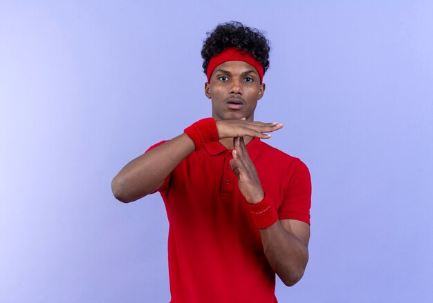 Looking at camera young afro-american sporty man wearing headband and wristband showing timeout gesture isolated on blue background