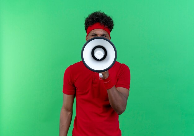 Looking at camera young afro-american sporty man wearing headband and wristband speaks on loudspeaker isolated on green background