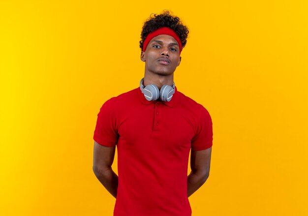 Looking at camera young afro-american sporty man wearing headband and wristband with headphones keeping hands behind back isolated on yellow background