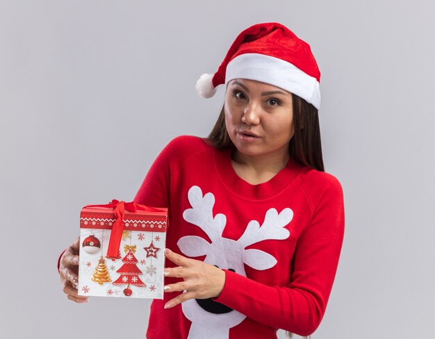 白い背景で隔離のギフトボックスを保持しているセーターとクリスマス帽子をかぶってカメラを見て若いアジアの女の子