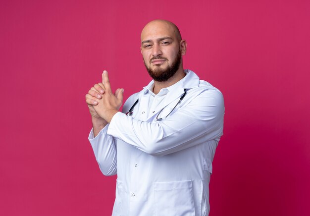 Looking at camera young bald male doctor wearing medical robe and stethoscope showing pistol gesture isolated on pink background