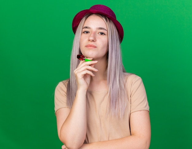 Looking at camera young beautiful girl wearing party hat holding party whistle putting hand under chin 