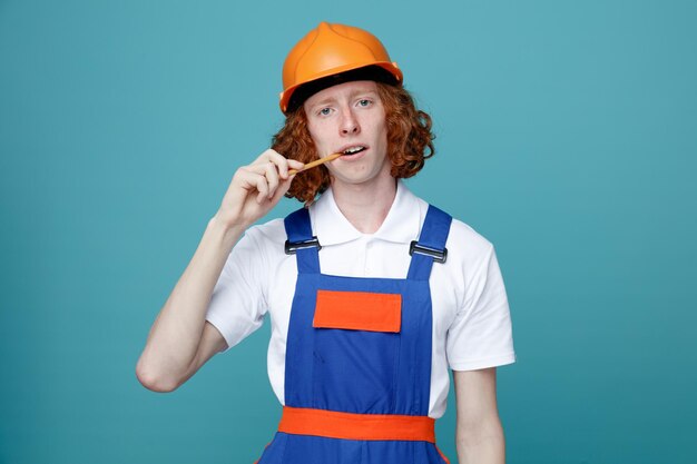 Looking at camera young builder man in uniform holding pencil in mouth isolated on blue background