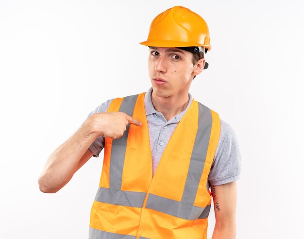 Looking at camera young builder man in uniform points at himself 