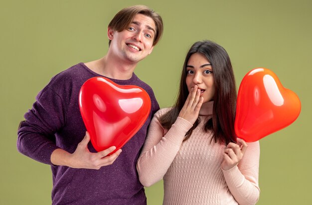 Looking camera young couple on valentines day holding heart balloons isolated on olive green background
